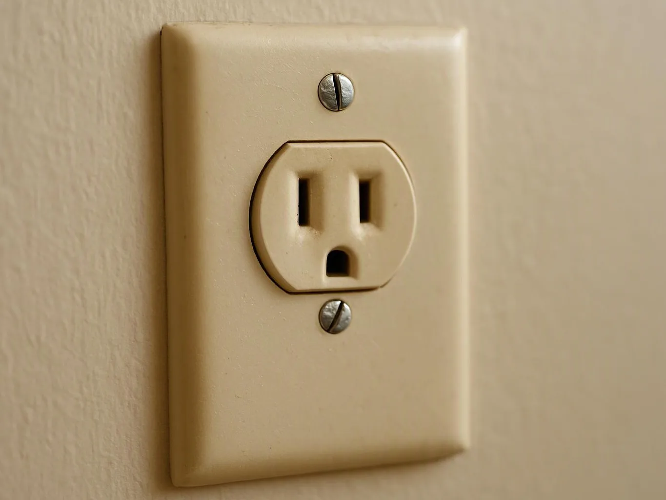 A beige wall outlet with two screws sits on a plain painted wall