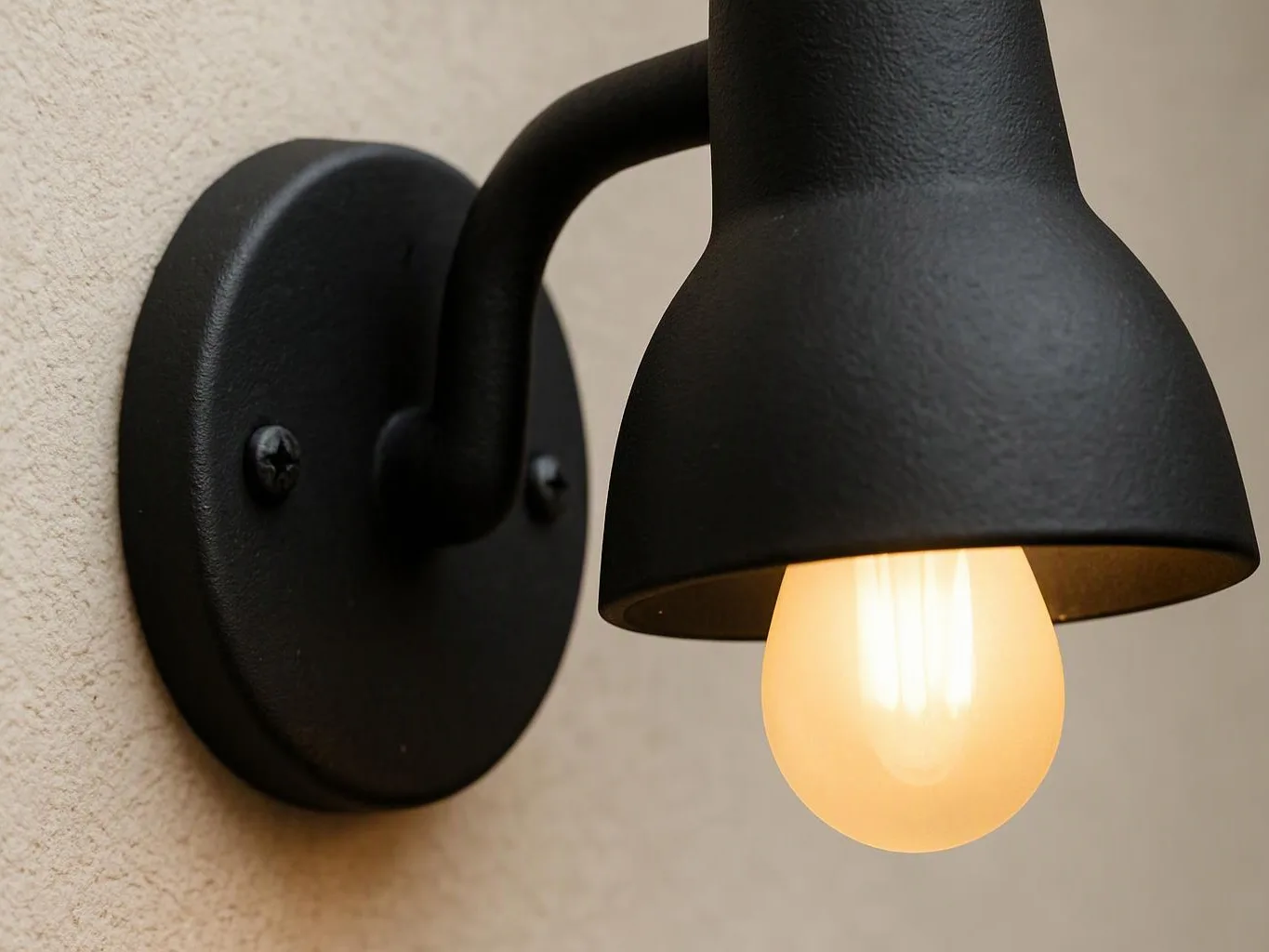 A black wall sconce with a glowing bulb hangs on a beige textured wall