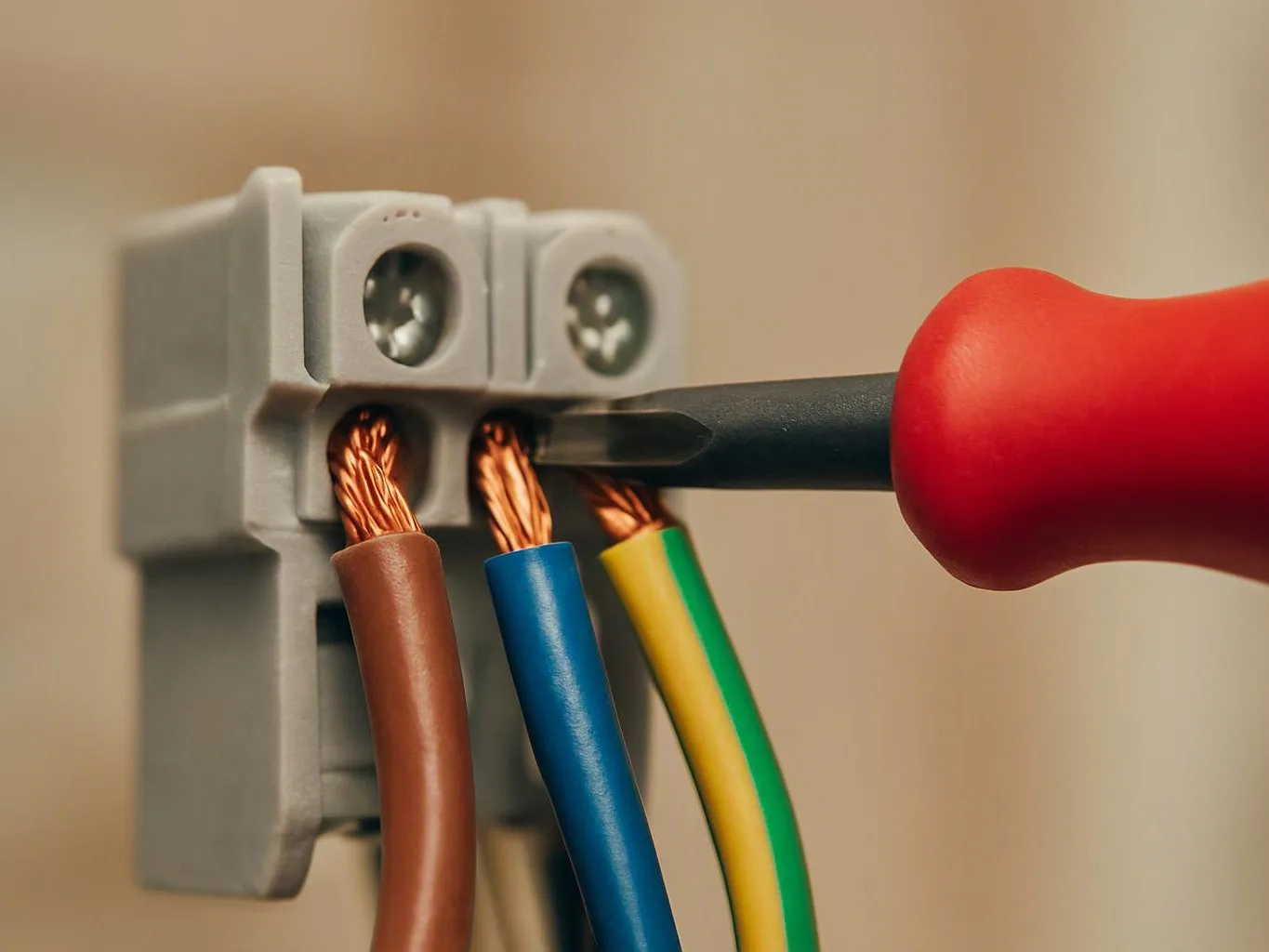 A screwdriver tightens a wire connector with three colored cables against a blurred beige background