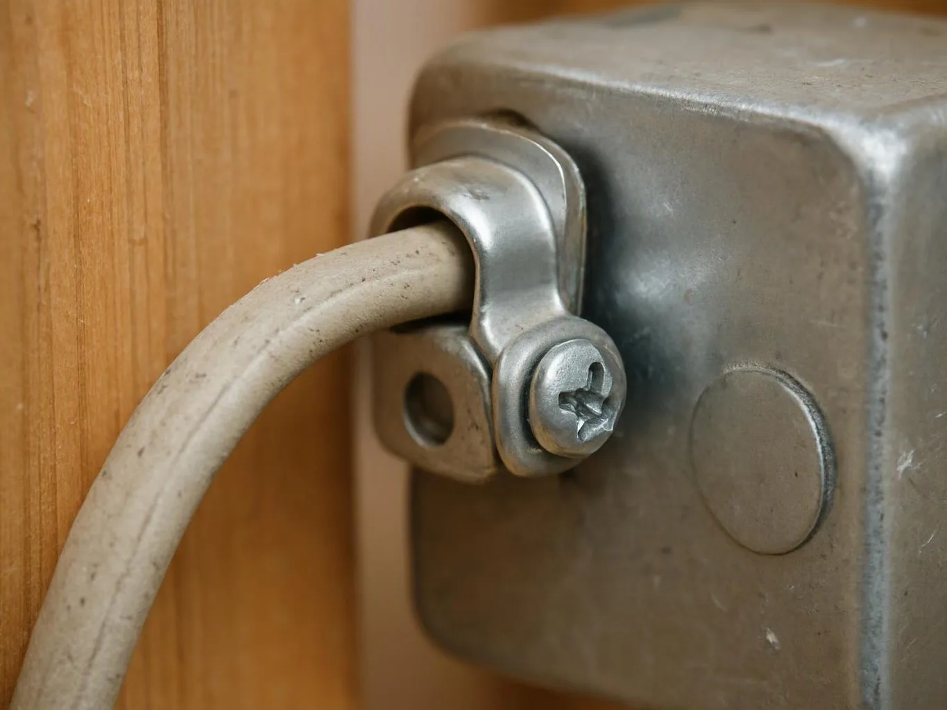 A white cable passes through a metal clamp on a gray electrical box beside a wooden panel